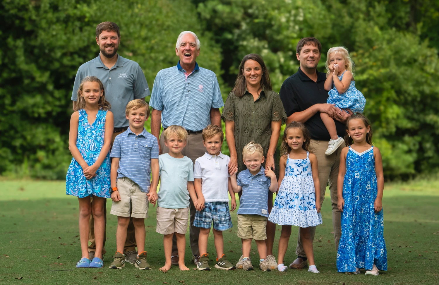 Our Story A large family portrait of three generations standing on a grassy lawn with a lush green background. The group includes three adult men, two adult women, and nine children of various ages. The adults are smiling, and the children are positioned in front and around them.
